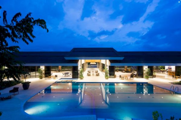 A luxurious modern building with large open spaces and a well-lit swimming pool at dusk, framed by a vibrant evening sky.