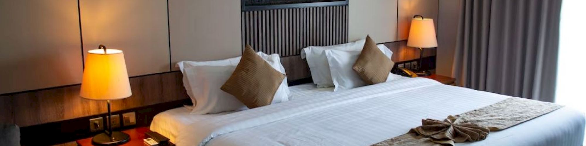 This image shows a neatly arranged hotel room with a large bed, decorative pillows, lamps on side tables, and a wall-mounted decorative piece.