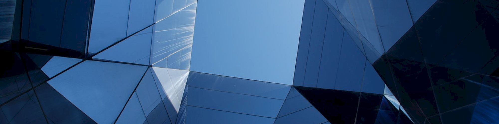 The image shows a geometric structure made of glass panels, forming an abstract and reflective pattern with a view of the sky at the center.