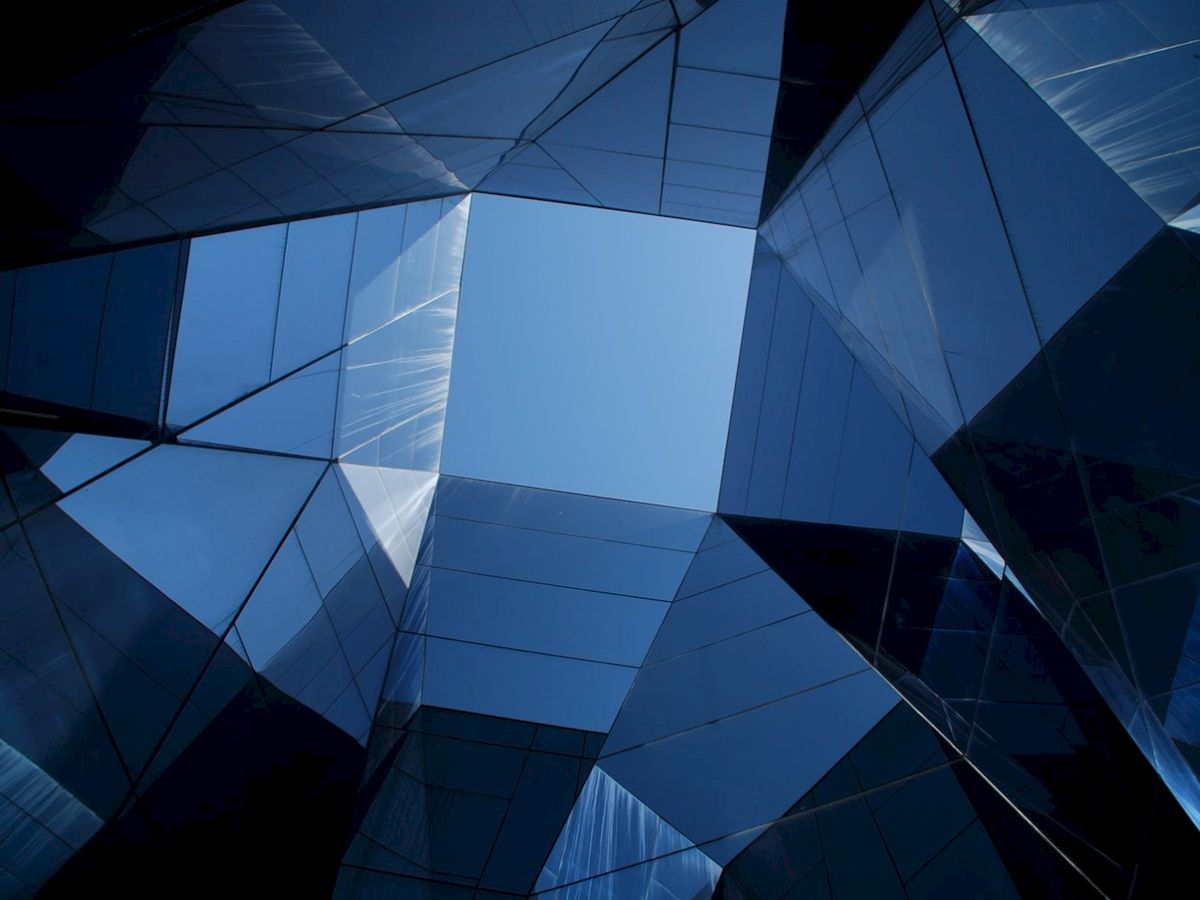 The image shows a geometric pattern formed by mirrored glass surfaces reflecting the sky, creating a modern architectural design with sharp angles.