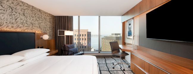 A modern hotel room with a large bed, a TV, a desk, a chair, and a window with a city view.