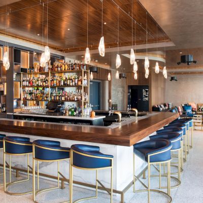 The image depicts a modern bar with high stools, a well-stocked bar counter, and hanging lights. The space appears to be well-lit and elegant.