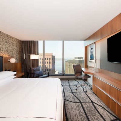 The image shows a modern hotel room with a large bed, wall-mounted TV, desk, chair, and a window with a cityscape view.