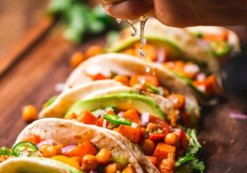 The image shows a hand squeezing lime juice over a row of tacos filled with chickpeas, diced tomatoes, avocado slices, and fresh herbs on a wooden board.