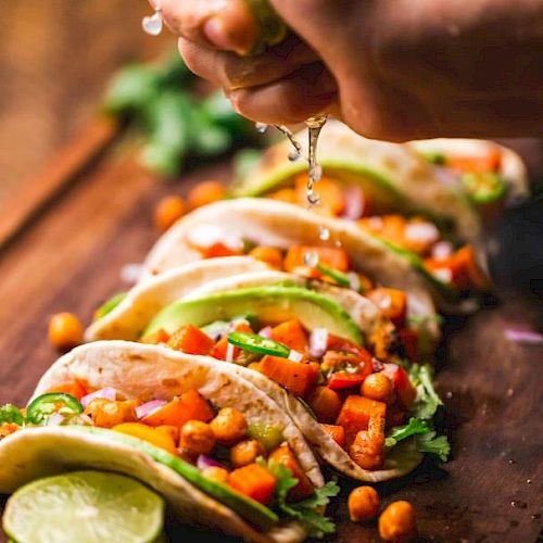 The image shows a hand squeezing lime juice over a row of tacos filled with chickpeas, diced tomatoes, avocado slices, and fresh herbs on a wooden board.