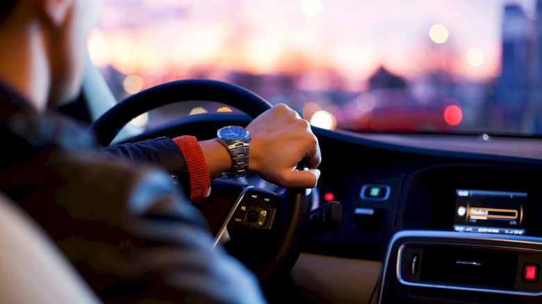 A person is driving a car during sunset with their left hand on the steering wheel and a view of the car's dashboard and instrument panel.
