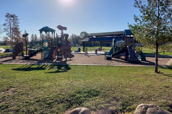 This image shows a playground with slides, climbing structures, and trees, set in a grassy area under a clear, sunny sky.