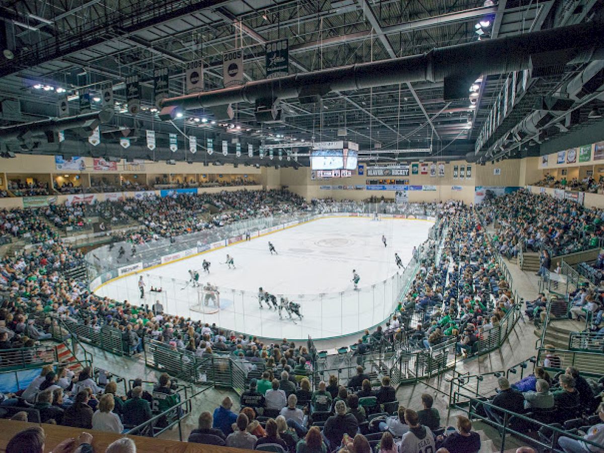 A crowded ice hockey arena with players on the rink and spectators filling the stands, all under bright lighting and a high ceiling.