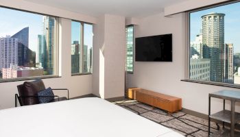 A modern hotel room with a city view, featuring large windows, a wall-mounted TV, a chair, and minimalistic furniture, showcasing an urban skyline.