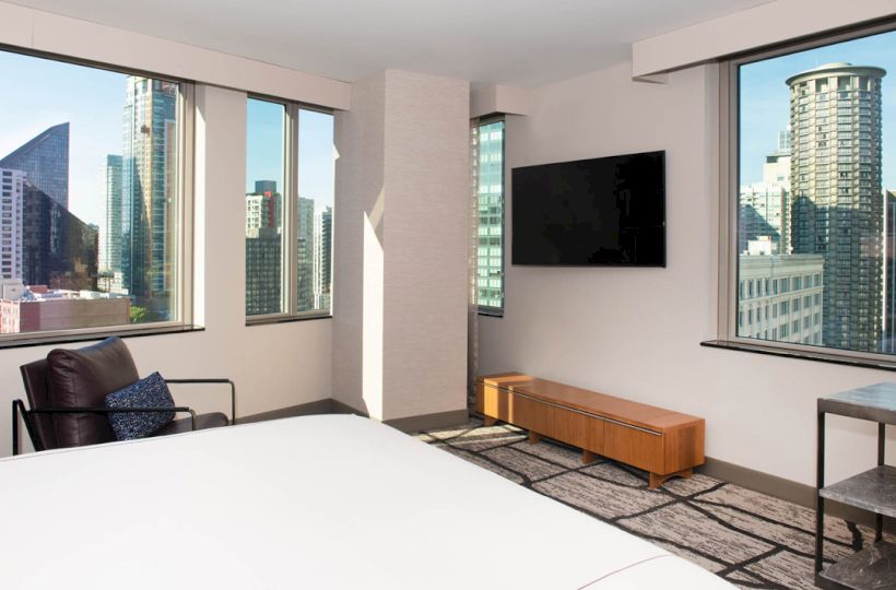 A modern hotel room with a city view, featuring large windows, a wall-mounted TV, a chair, and minimalistic furniture, showcasing an urban skyline.