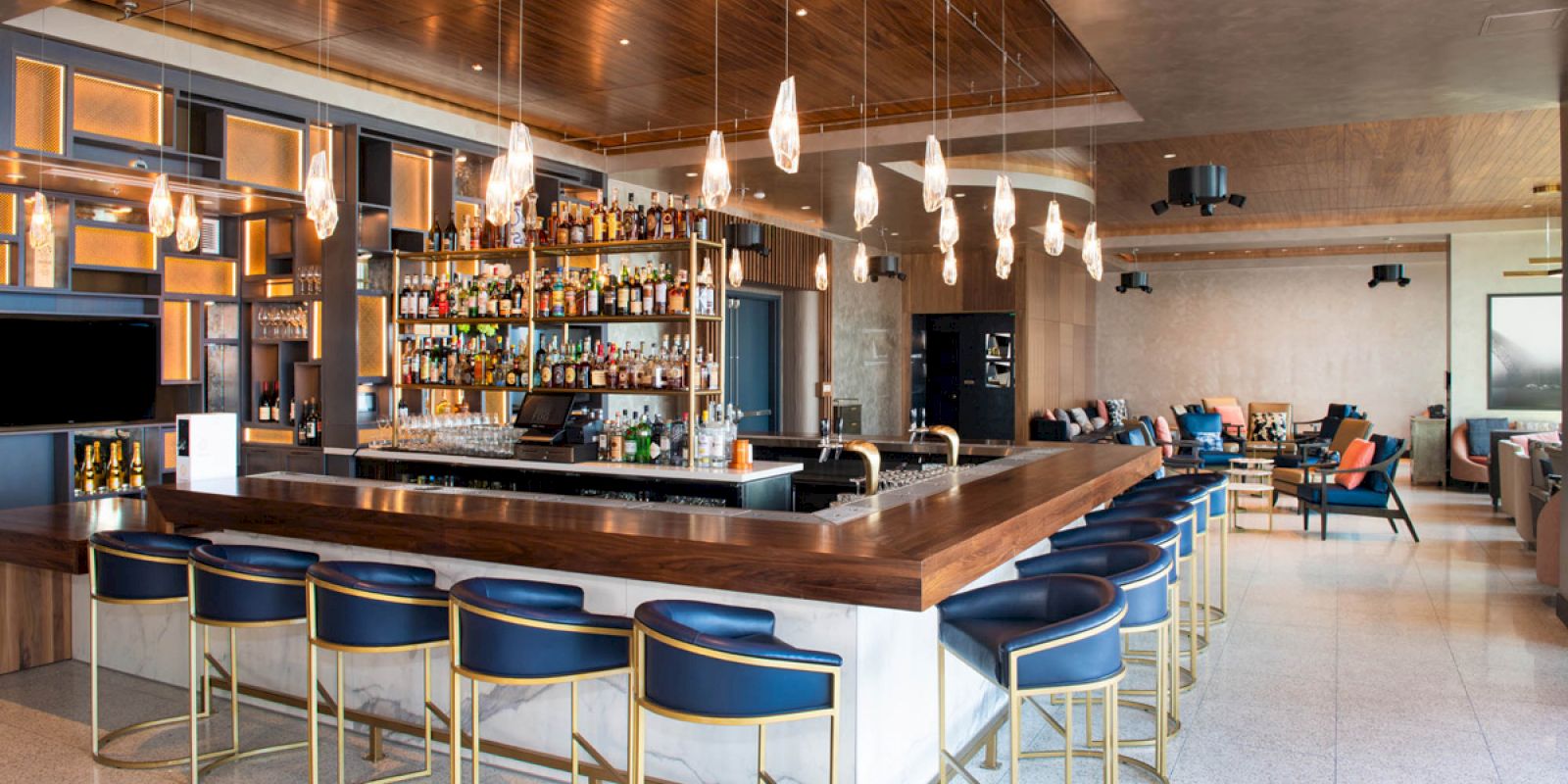 A modern bar with blue cushioned stools, a well-stocked liquor shelf, elegant pendant lights, and a cozy lounge area in the background.