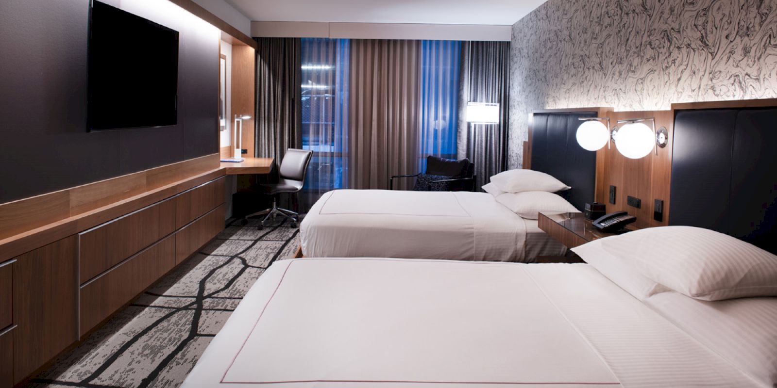 A modern hotel room features two neatly made beds, a large flat-screen TV, a desk with a chair, and a patterned carpet, all lit by stylish lamps.