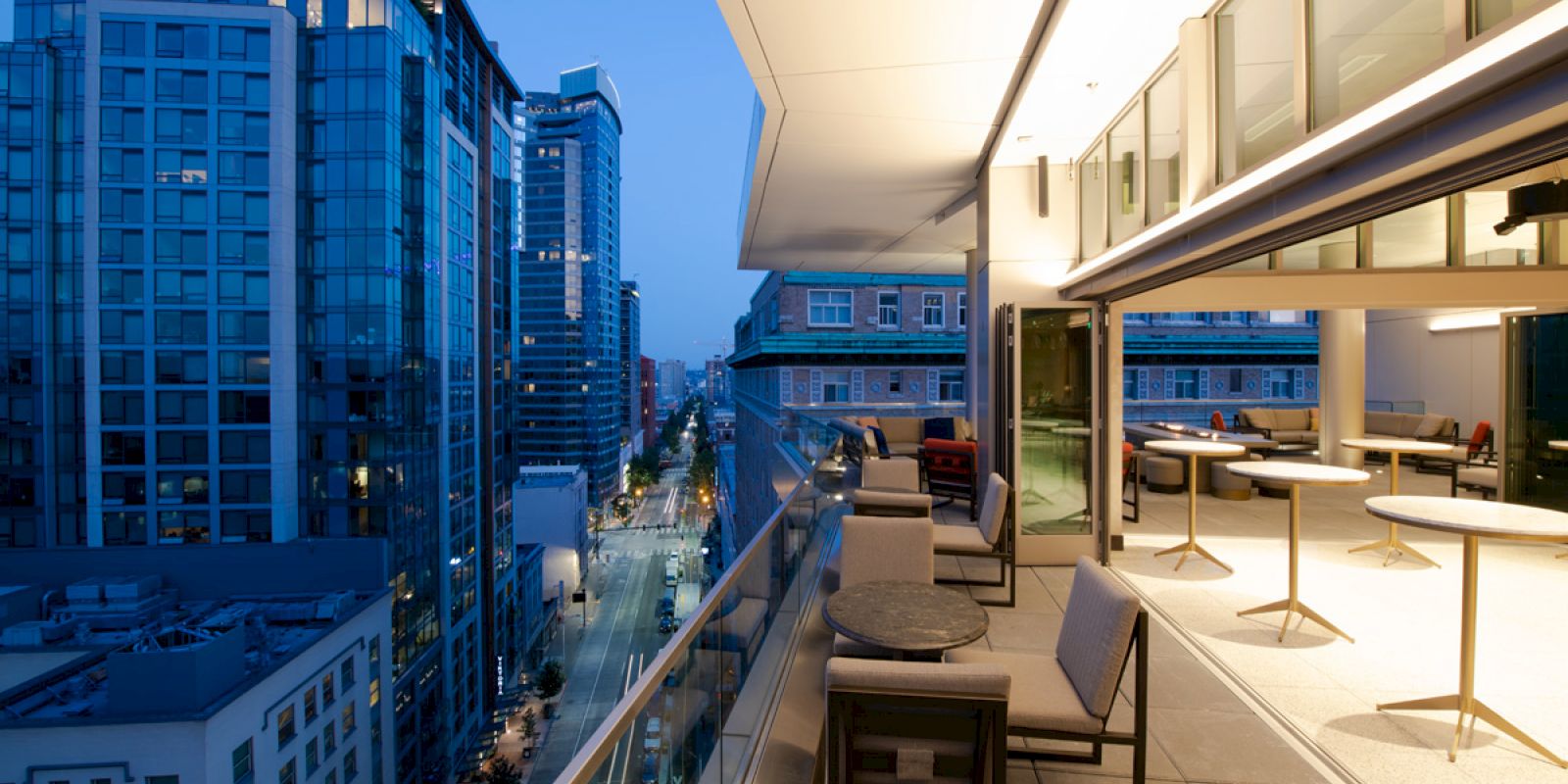 A modern urban balcony at dusk features comfortable seating and tables, overlooking a cityscape with high-rise buildings and a well-lit street.