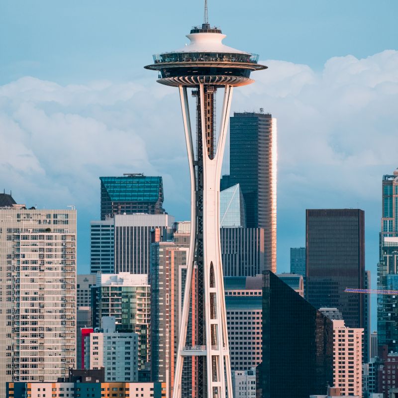 The image shows a skyline with a tall structure resembling the Space Needle in the foreground, surrounded by various modern buildings.