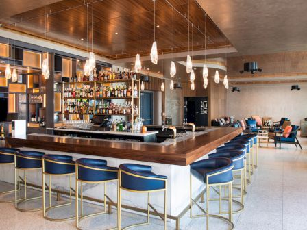 This image features a modern bar with blue bar stools, hanging lights, and a well-stocked shelf of liquor bottles. The interior is stylish and inviting.
