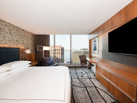 A modern hotel room with a large bed, desk, chair, flat-screen TV, and a window with a city view. The decor is sleek and contemporary.