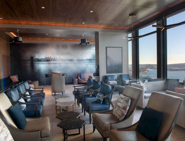 The image shows an upscale lounge area with modern seating, large windows offering a scenic waterfront view, and contemporary decor.