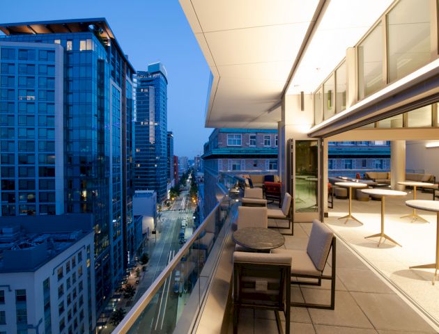 The image shows a modern balcony at dusk with seating and tables, overlooking a city street lined with tall buildings and glowing lights.