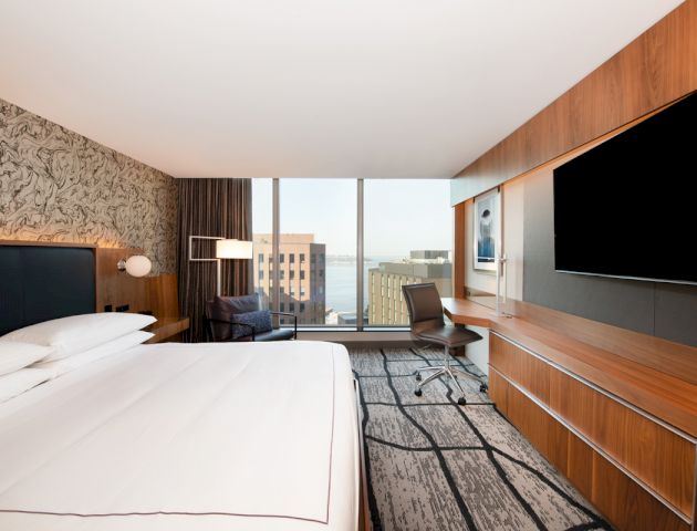 This image shows a modern hotel room with a large bed, wall-mounted TV, desk, and a view of tall buildings through the window.