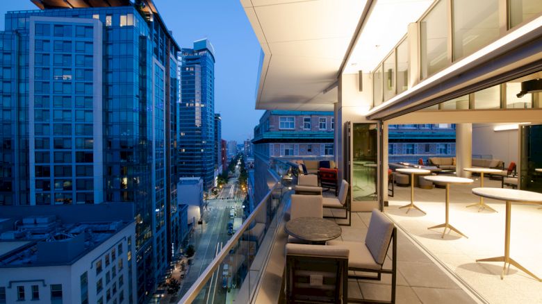 A modern outdoor balcony with seating and tables overlooks a cityscape with high-rise buildings and a street below, lit up in the evening.