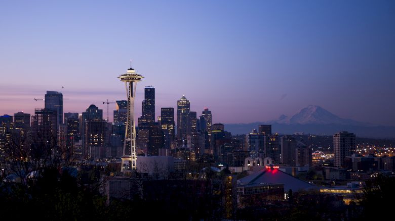 The image features the Seattle skyline at dusk, showcasing the Space Needle and Mount Rainier in the background, with the city lights starting to illuminate.