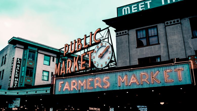 The image shows the iconic entrance sign of a public farmers market, with neon lights and a clock, surrounded by various buildings and shops.