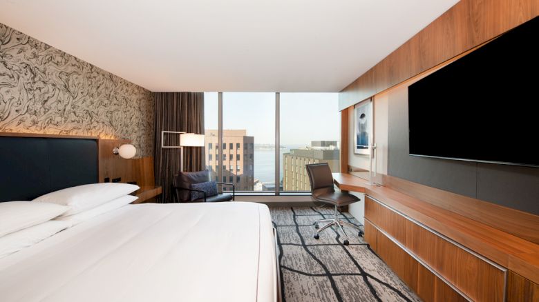 This image shows a modern hotel room with a large bed, a desk and chair, a flat-screen TV, and a view of city buildings through a large window.