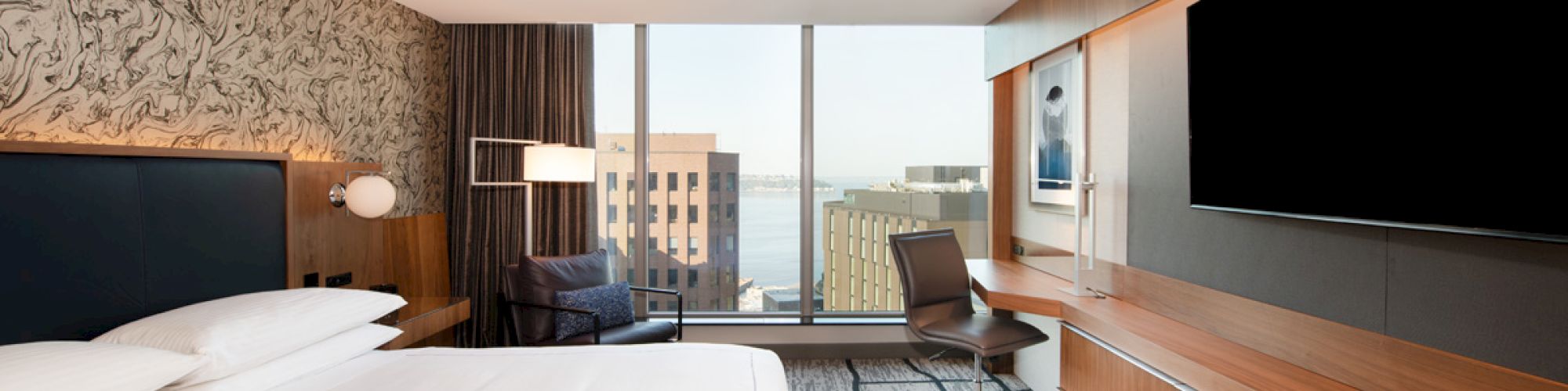A modern hotel room features a large bed, a desk with a chair, a wall-mounted TV, and a window with a city and water view.
